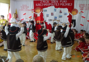 Na tle dekoracji z mapą Polski i dziećmi z chorągiewkami przedszkolaki przebrane za górali tańczą z ciupagami.
